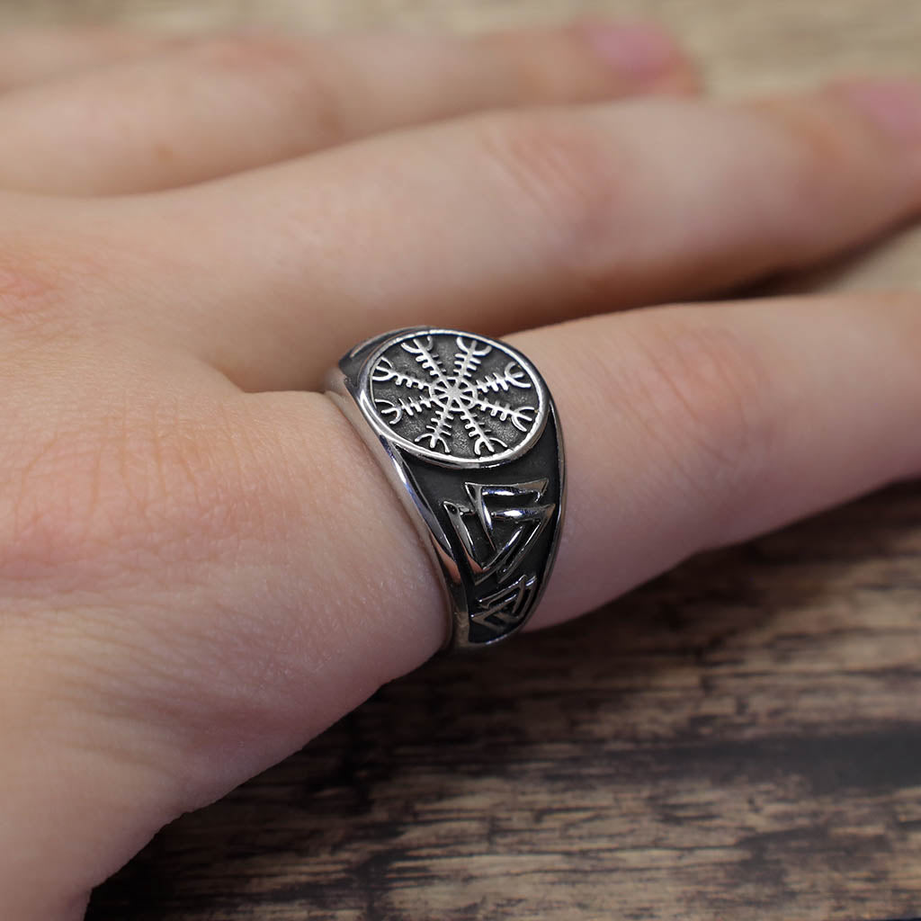 A silver signet ring featuring an engraved Vegvisir symbol on the face and Valknut motifs along the band, shown worn on a finger against a wooden surface.