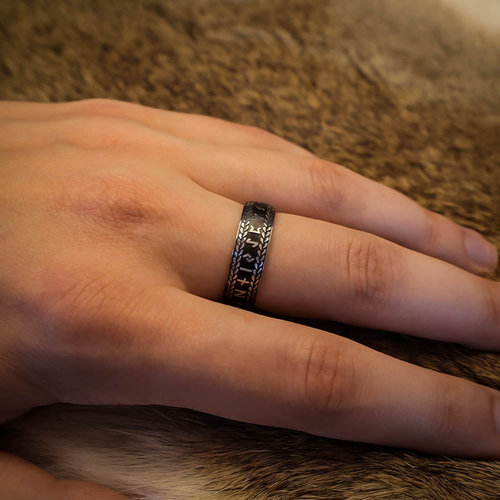 A hand wearing a black and silver ring engraved with Norse runes and a braided border, resting on a textured fur surface.