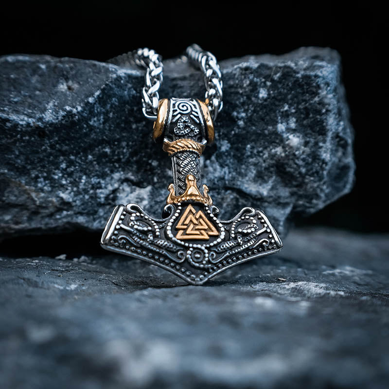 A stainless steel Thor's Hammer necklace with intricate engravings and a Valknut rune at the center, displayed against a rocky background.