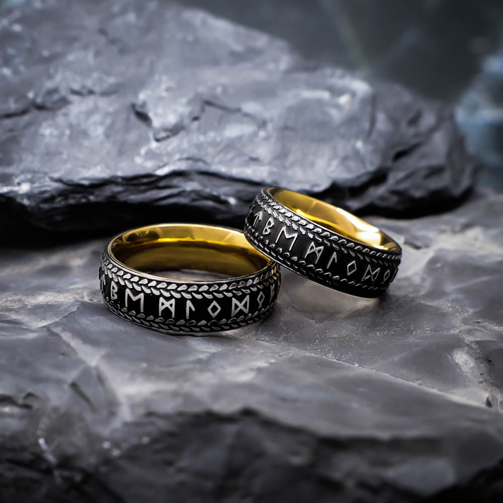 Two gold-lined black rings engraved with Norse runes and bordered by a braided pattern, displayed on dark textured stones in a close-up setting.