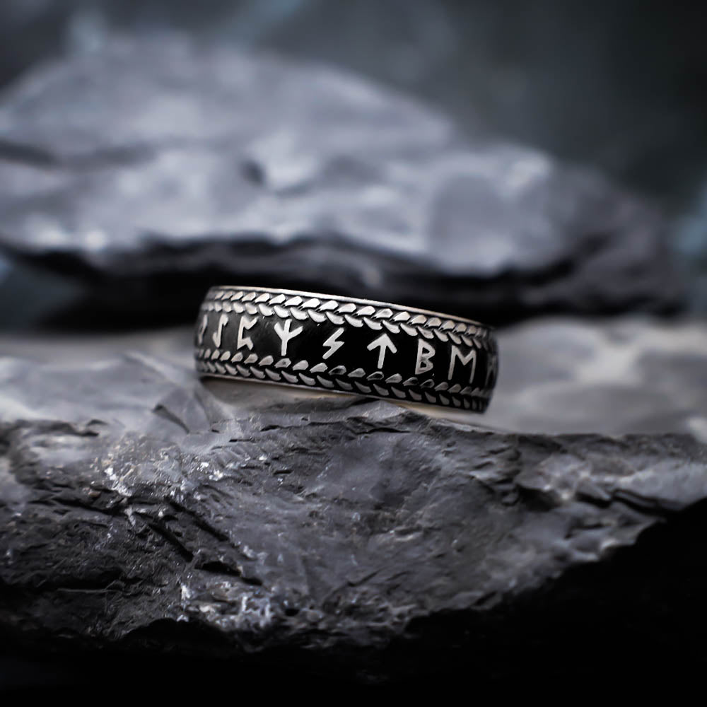 A black and silver ring engraved with Norse runes and framed by a braided border, resting on dark textured stones in a moody close-up composition.