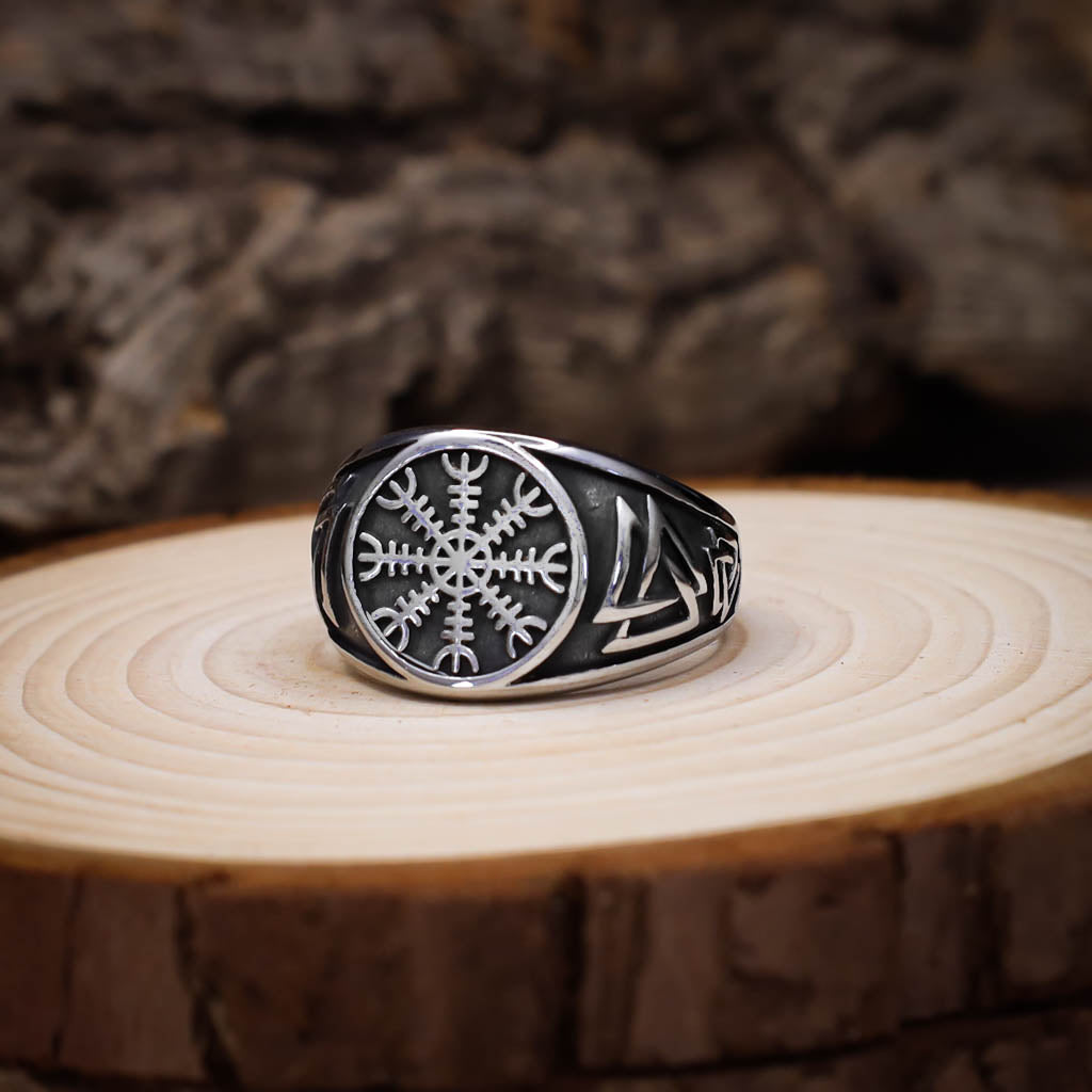 A silver signet ring featuring an engraved Vegvisir symbol on the face and Valknut motifs on the sides, displayed on a circular cut of light wood with a rustic, textured background.