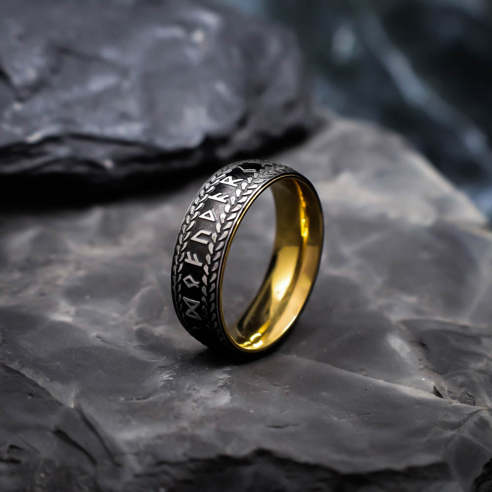 A close-up of a gold-lined black ring engraved with Norse runes and bordered by a braided pattern, displayed upright on dark textured stone.