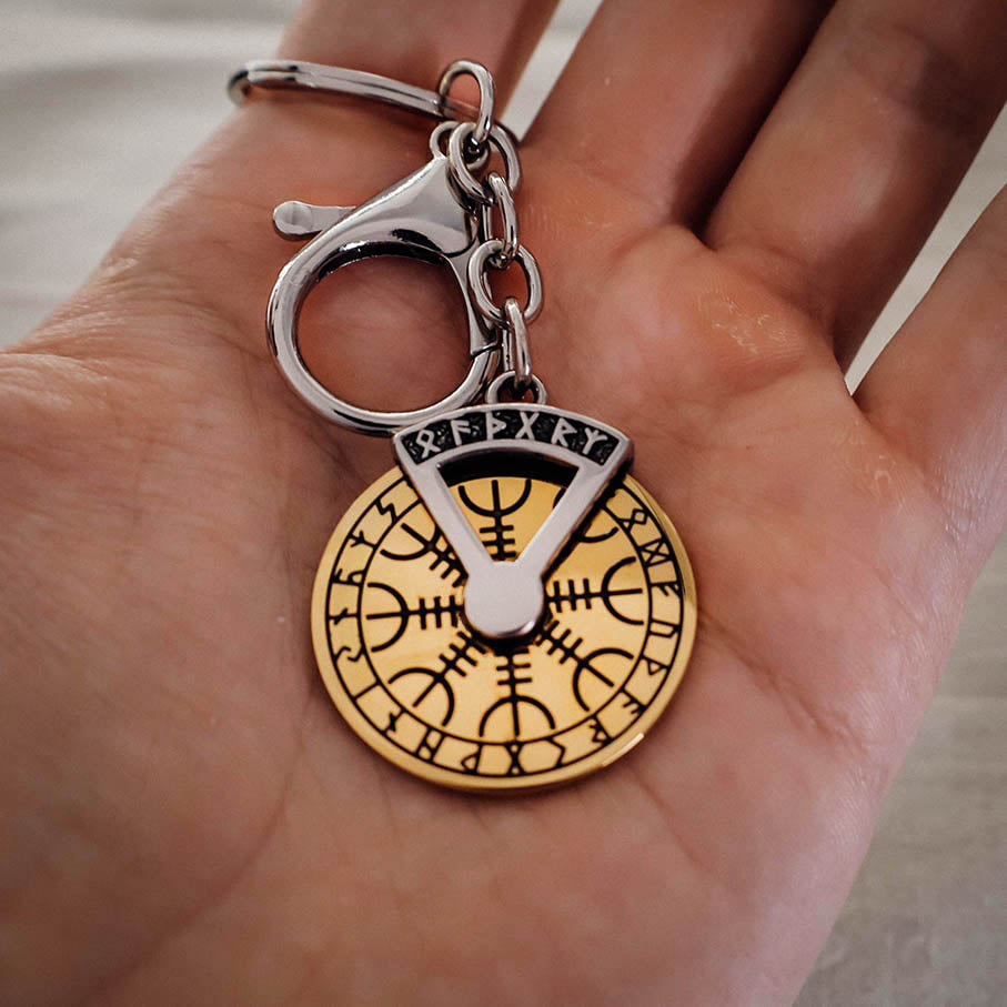 A hand holding a metallic keychain featuring a gold circular pendant engraved with Norse runes and a black symbol design, attached to a silver clasp and chain.