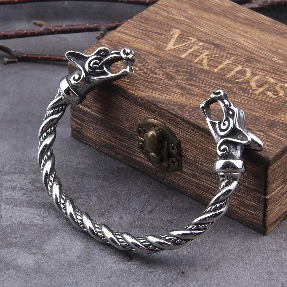 A twisted stainless steel bracelet with intricately detailed wolf head terminals, displayed beside a wooden box engraved with the word “Vikings” on a gray stone surface.