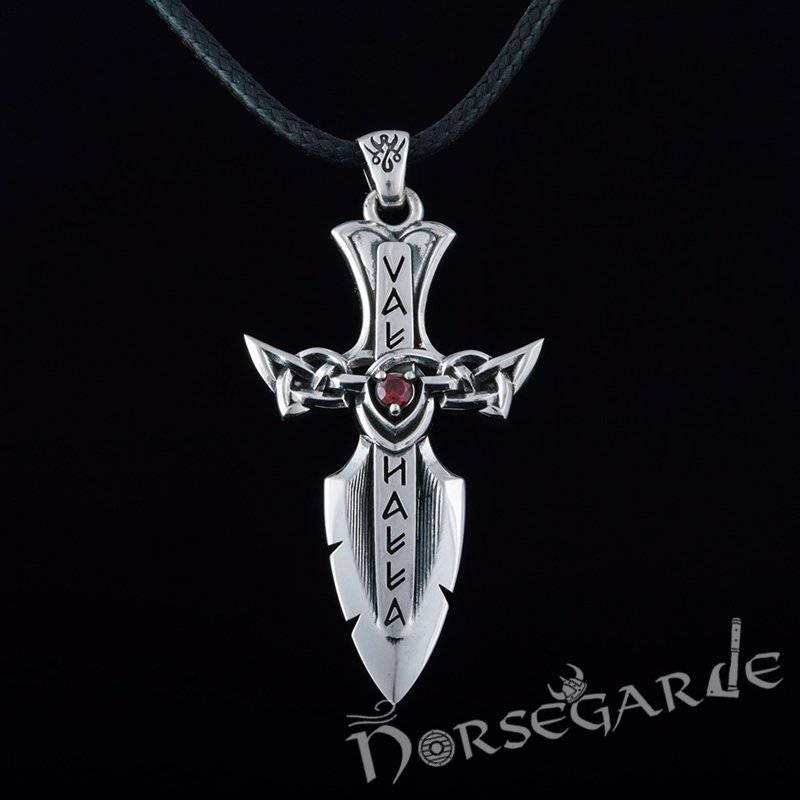 A silver Valhalla-themed dagger pendant with runic-style lettering reading “VAL HALLA,” intricate knotwork forming the crossguard, and a red gemstone set at the center, hanging from a black braided cord against a dark background