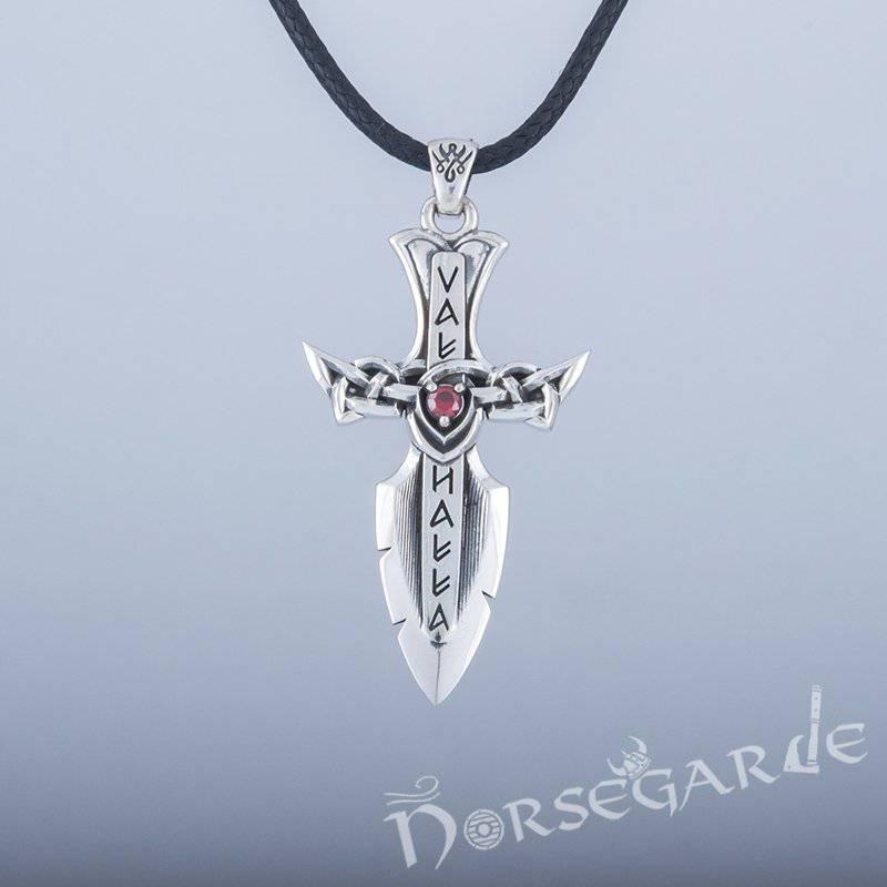 A silver Valhalla-themed dagger pendant with runic-style lettering and a red gemstone at the center of an intricate knotwork crossguard, hanging from a black braided cord against a light background