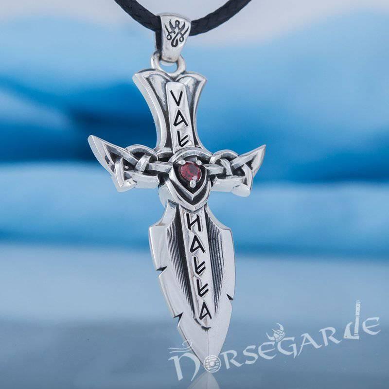 A silver Valhalla-themed dagger pendant with runic-style lettering, intricate knotwork forming the crossguard, and a red gemstone at the center, shown hanging from a black braided cord against a soft blue background