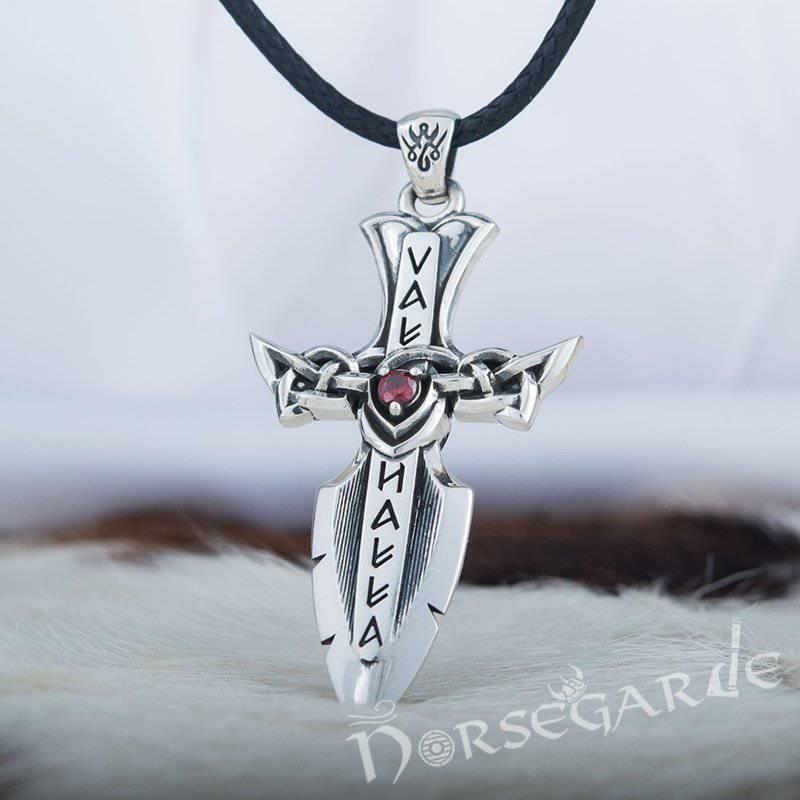 A silver Valhalla-themed dagger pendant with runic-style lettering, intricate knotwork forming the crossguard, and a red gemstone at the center, hanging from a black braided cord and displayed against a soft white and fur-textured background
