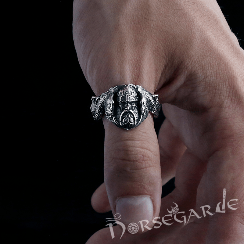 A silver ring featuring a detailed sculpted Norse warrior’s face flanked by raven wings, worn on a person’s finger against a dark background.