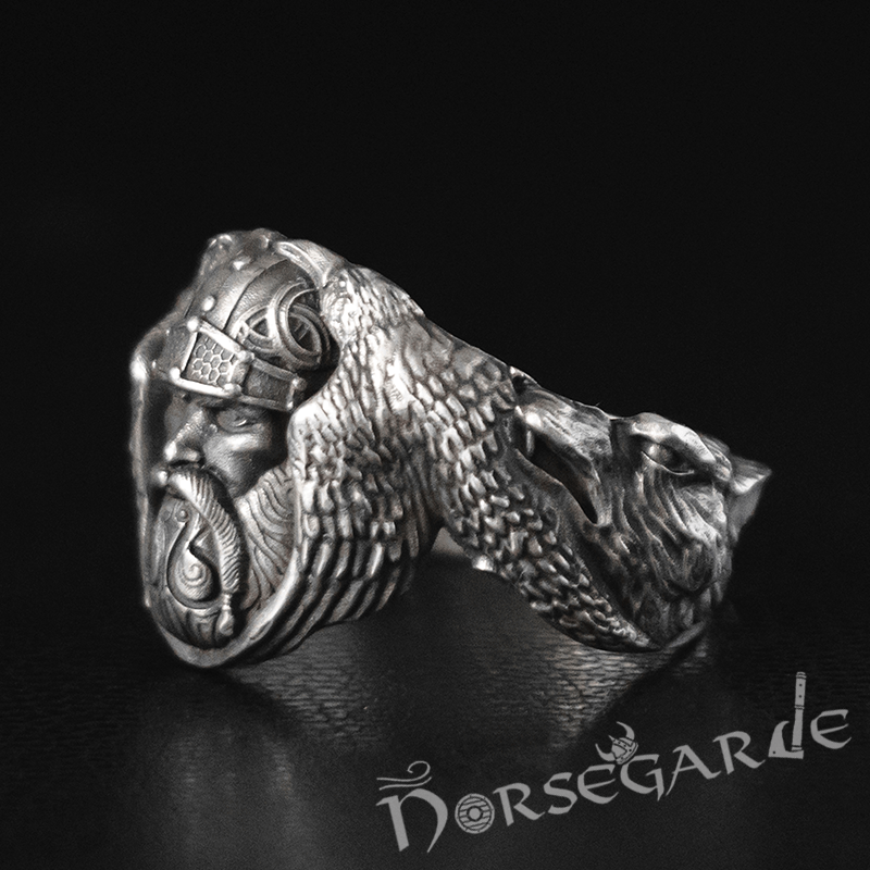 A detailed silver ring featuring a sculpted Norse figure wearing a helmet, flanked by textured raven wings that curve into a snarling wolf’s head, displayed on a dark surface with the Norsegarde logo below.