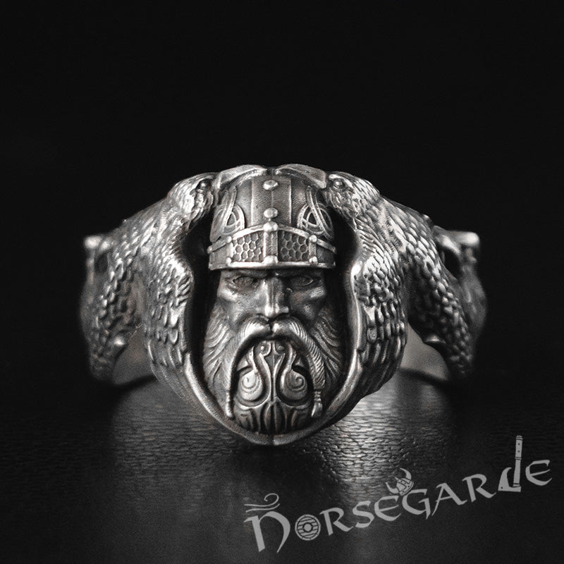 A detailed silver ring featuring a sculpted Norse warrior face wearing a horned helmet, flanked by two ravens, displayed against a dark background with the Norsegarde logo below.