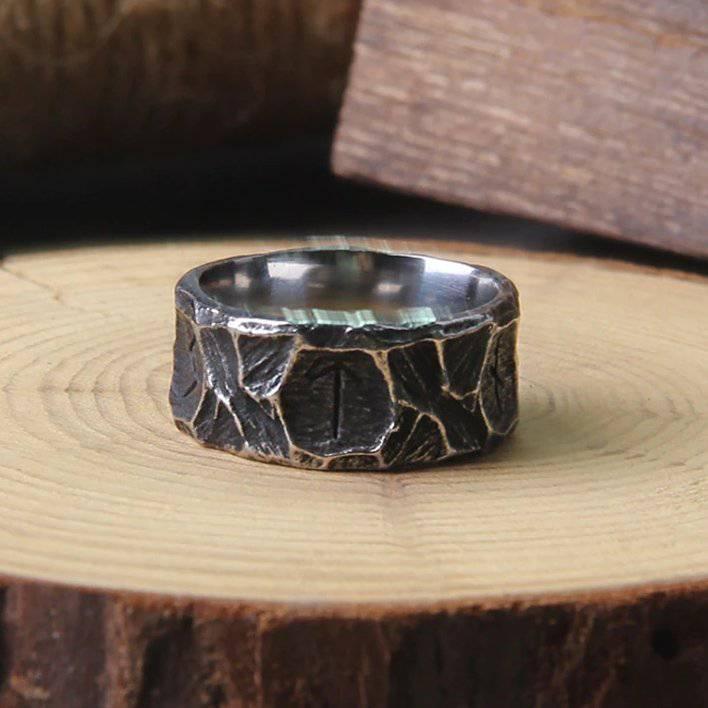A Viking-inspired stainless steel ring with Elder Futhark runes etched into a hammered texture surface, displayed on a wooden platform.