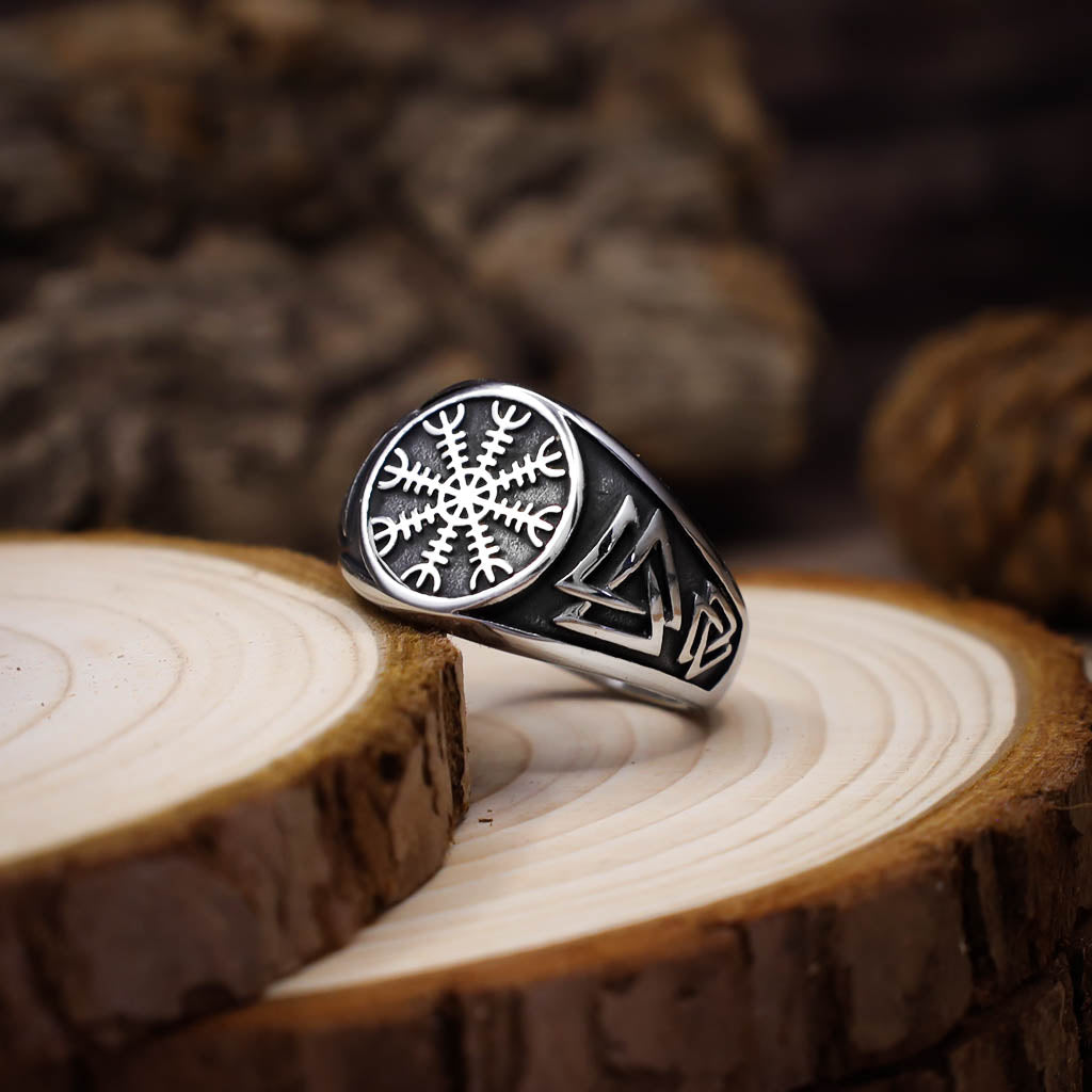 Aa angled up silver signet ring featuring an engraved Vegvisir symbol on the face and Valknut motifs along the band, displayed on stacked slices of light wood with a rustic, blurred background.