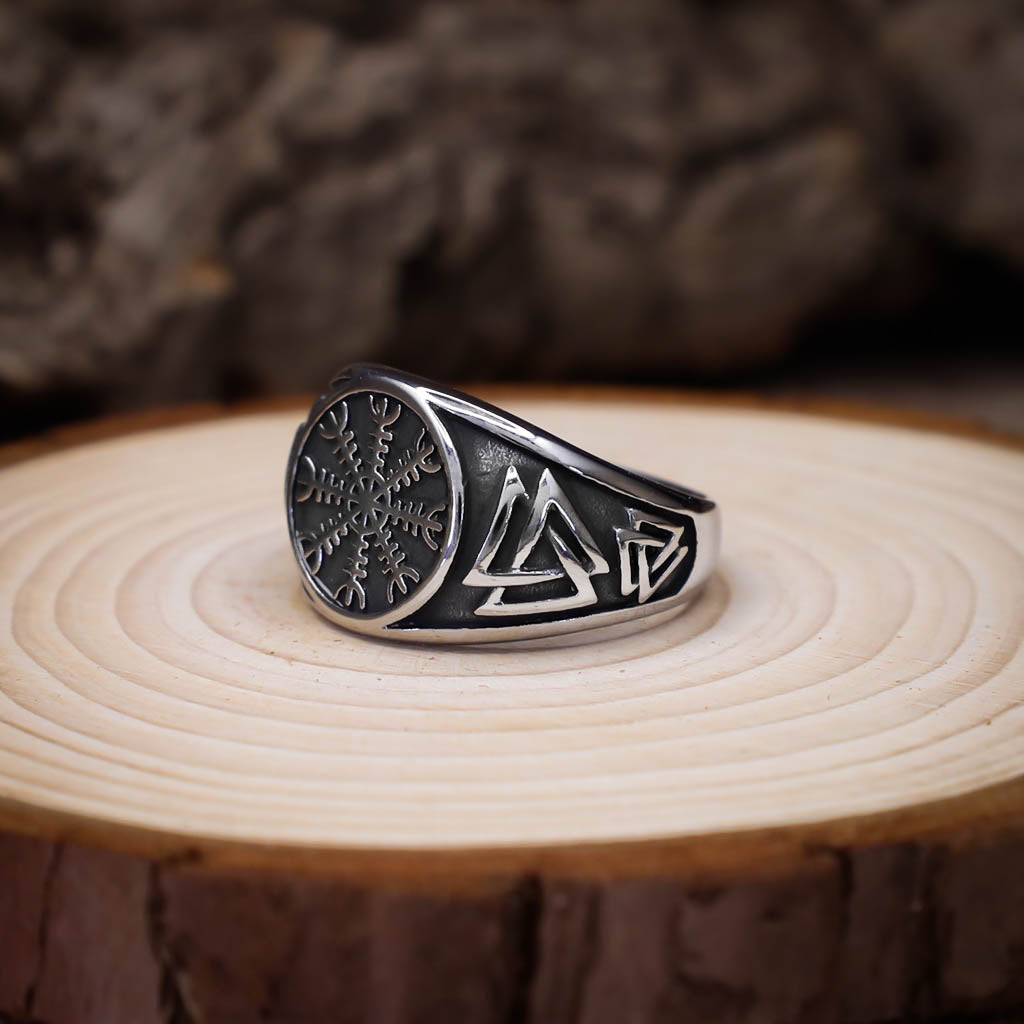 A silver signet ring featuring an engraved Vegvisir symbol on the face and Valknut designs along the side of the band, displayed on a circular slice of light wood with a rustic background.