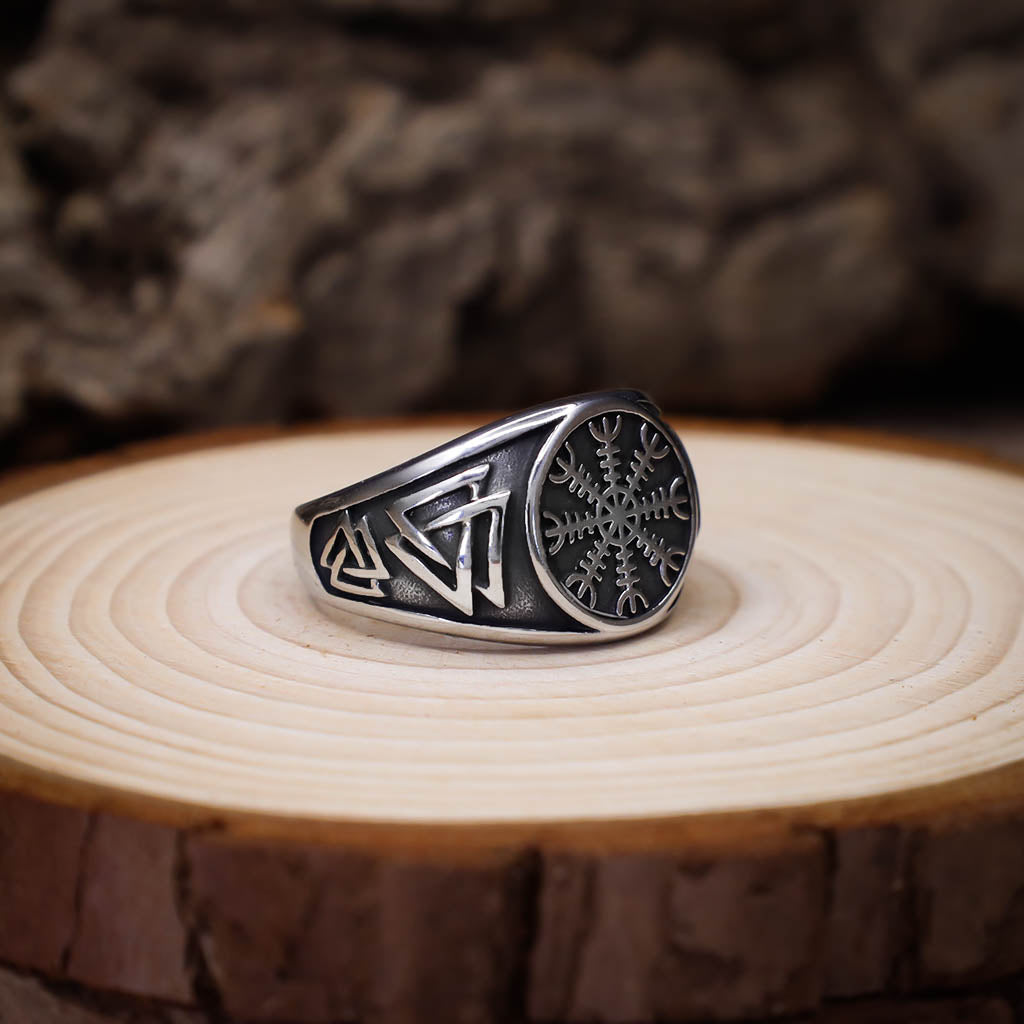 A silver signet ring featuring an engraved Vegvisir symbol on the face and multiple Valknut designs along the band, displayed on a circular slice of light wood with a rustic, textured background.