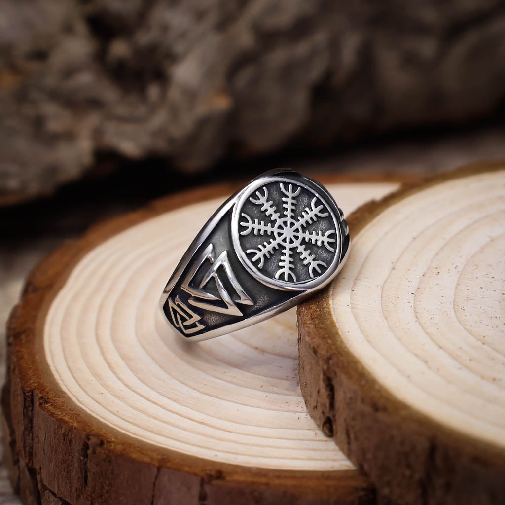 A silver signet ring featuring an engraved Vegvisir symbol on the face and Valknut designs on the sides, displayed on stacked slices of light wood with a rustic background.