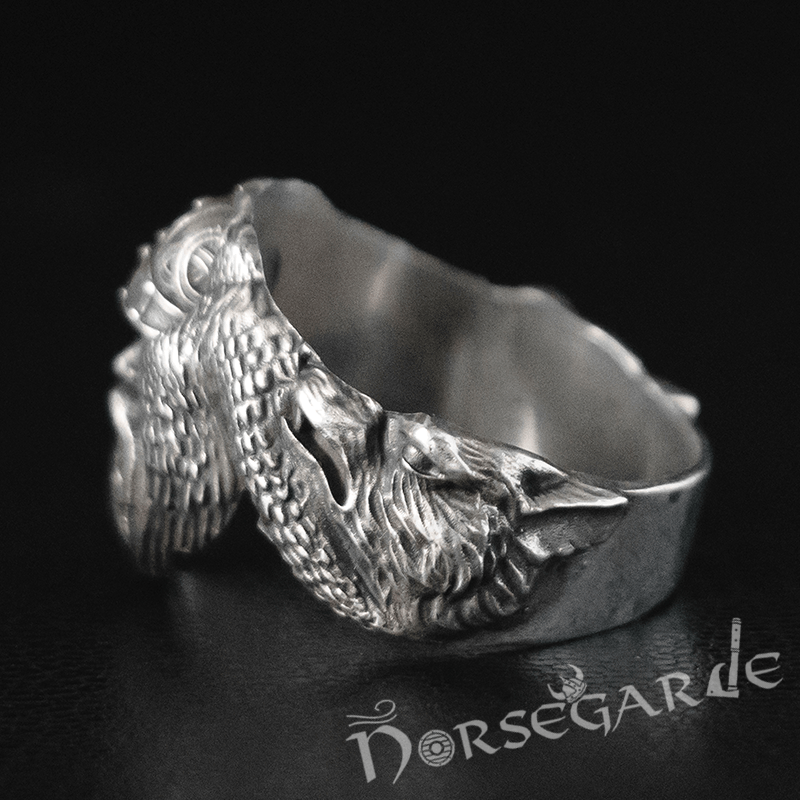 A silver ring featuring a sculpted wolf head merging into detailed raven wings, with Norse-inspired engraving near the band, displayed on a dark surface.