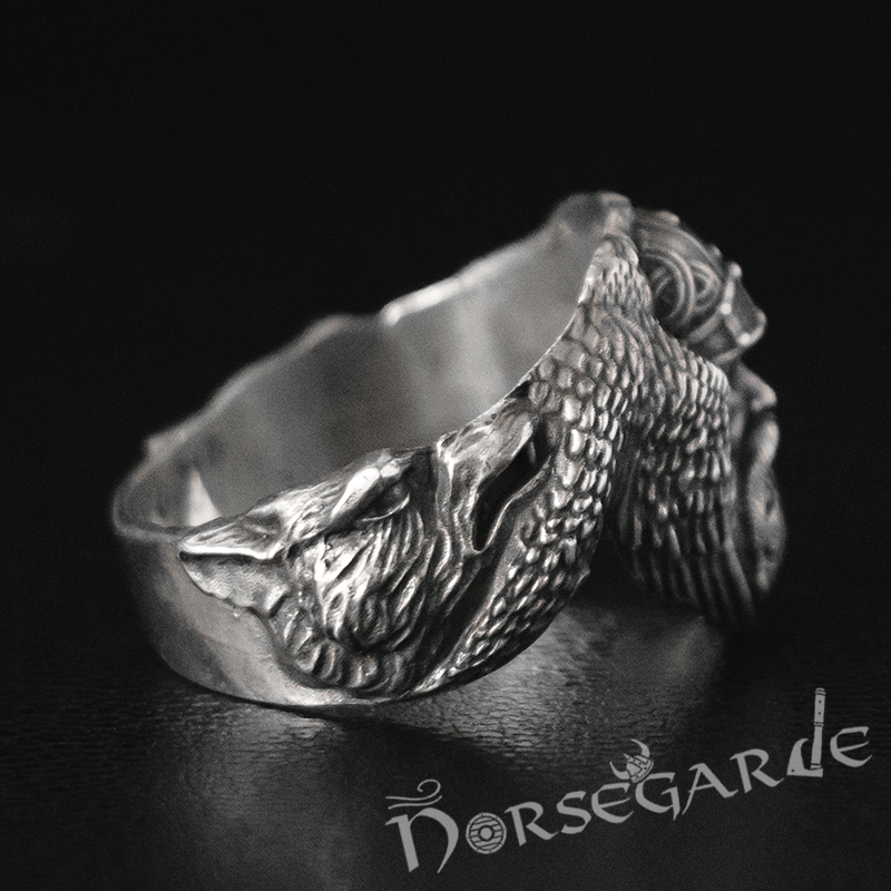 A silver ring featuring a detailed sculpted wolf head merging into textured raven wings, displayed on a dark surface.