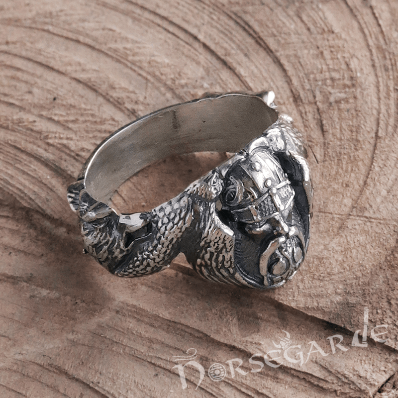 A handcrafted silver ring featuring a detailed Norse warrior’s face with a helmet, flanked by textured raven wings that transition into a wolf’s head, displayed on a carved wooden surface.