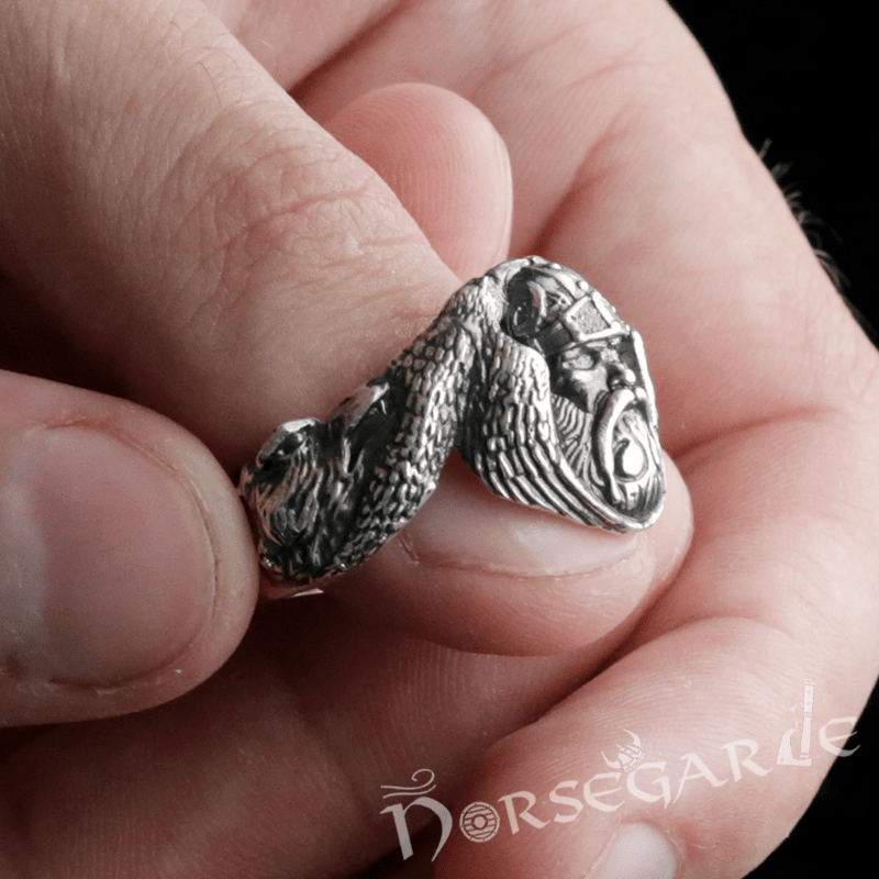 A detailed silver ring featuring a sculpted Norse warrior’s face and flowing raven wings, held between two fingers.